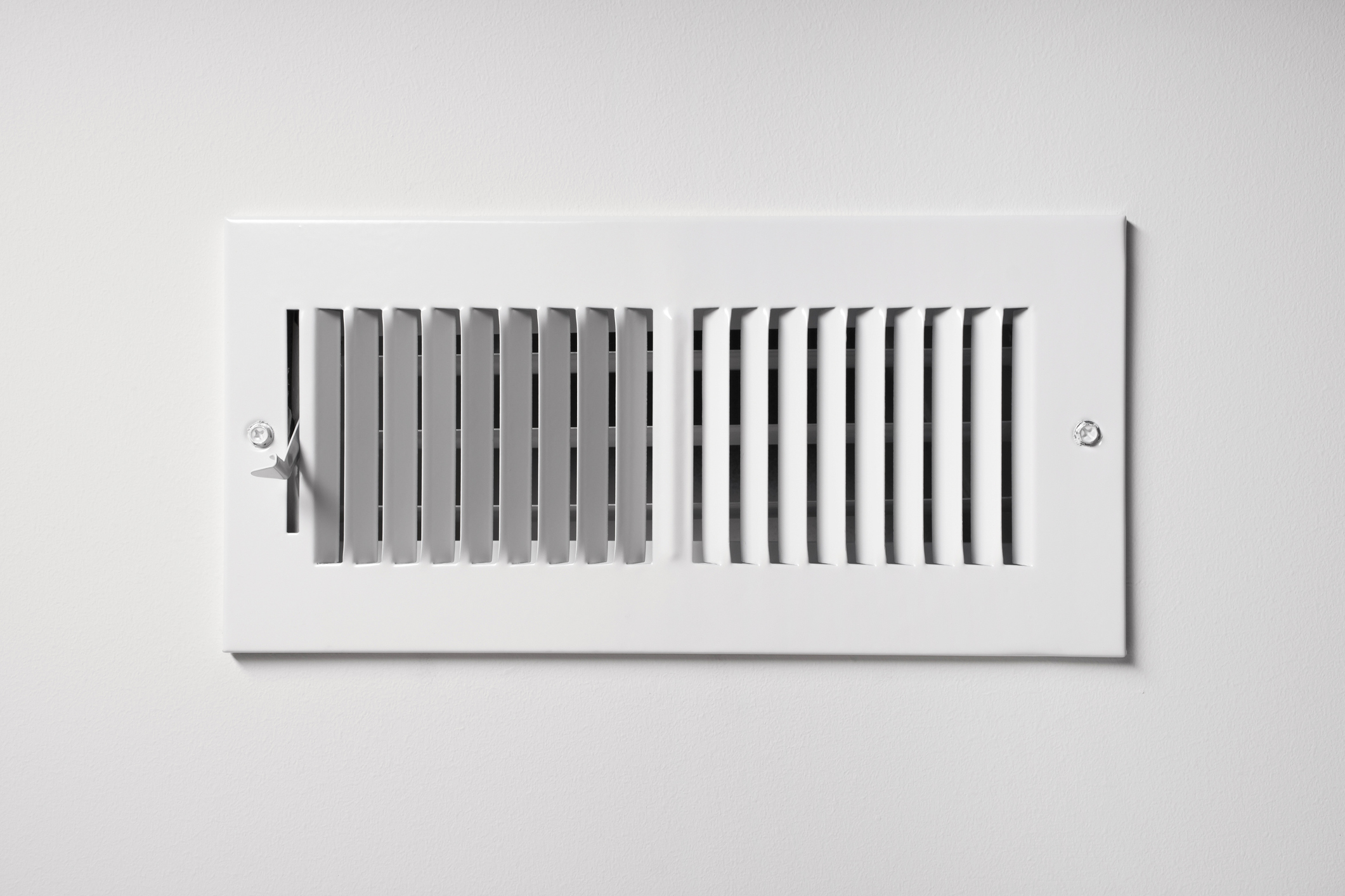 A heating/cooling vent register on the white wall of a home, with open/close lever.
