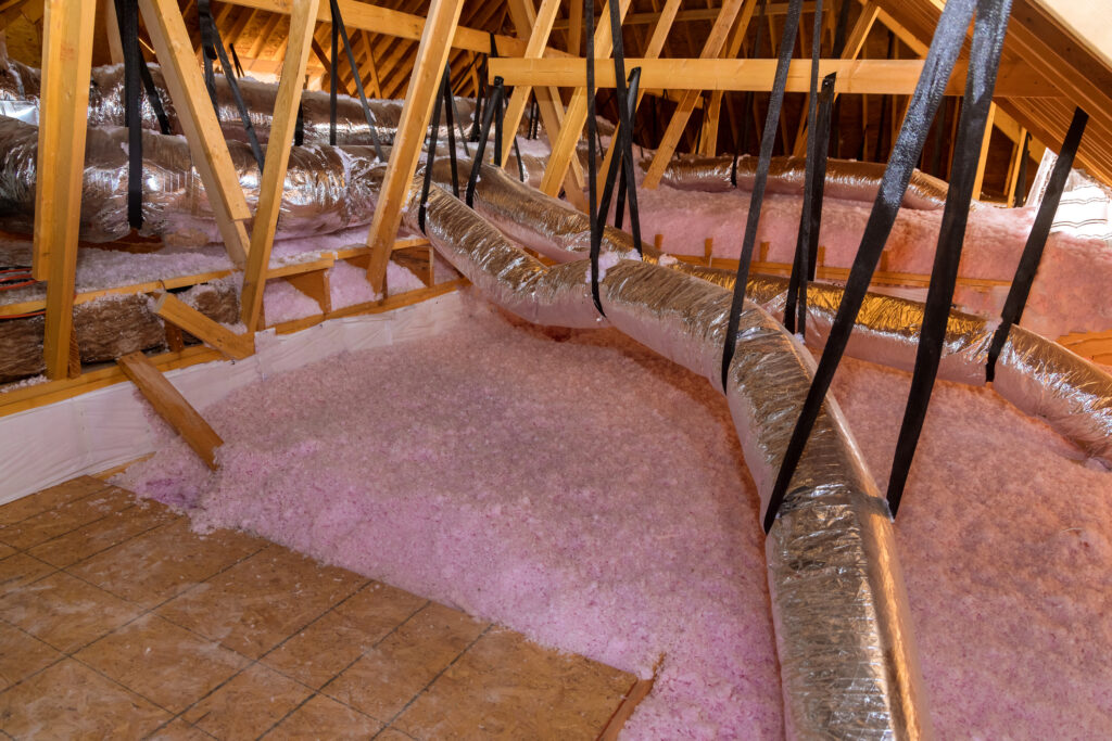 Ductwork installed in an unfinished attic with pink loose-fill insulation all over the floor.